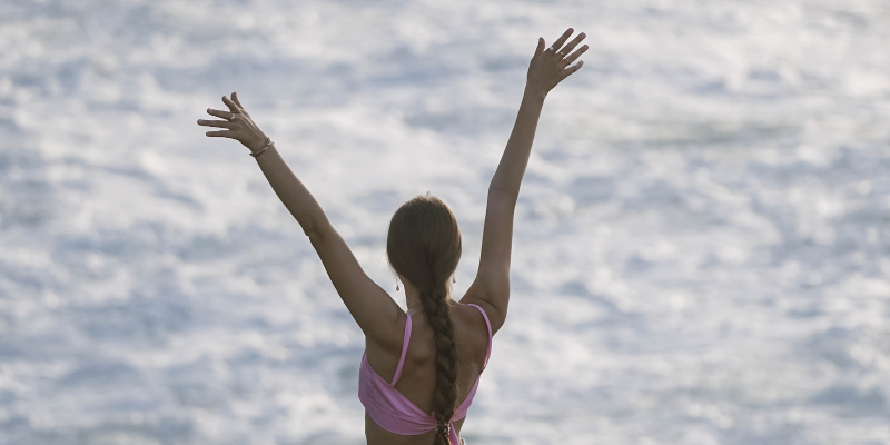 Young woman raises her hands in the air feeling pain free while looking at the ocean